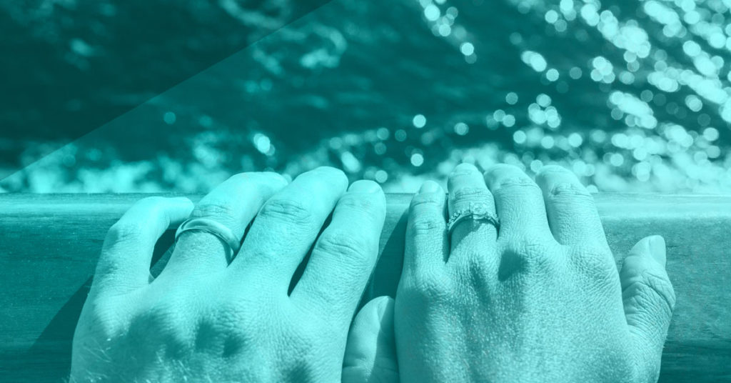 Hands wearing left finger wedding rings on the edge of a boat overlooking blue water