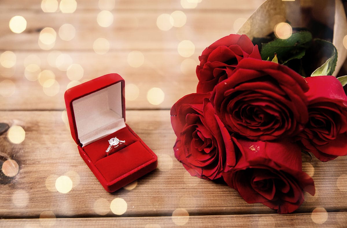 diamond ring in a box next to a bouquet of red roses on a wooden background