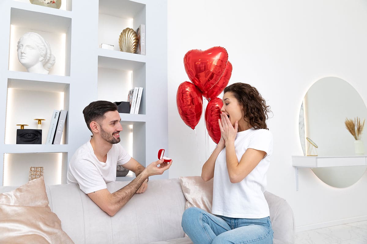 woman on couch with heart balloons smiling as man proposes